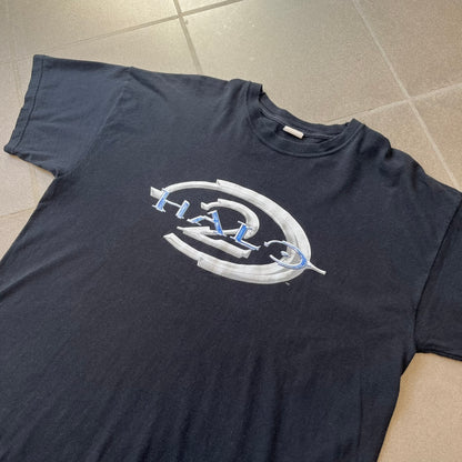 Halo 2 Xbox 2004 Black Tee is a black promotional T-shirt featuring a large white and blue Halo 2 graphic on the front. The upper back has a green original Xbox logo positioned below the collar.