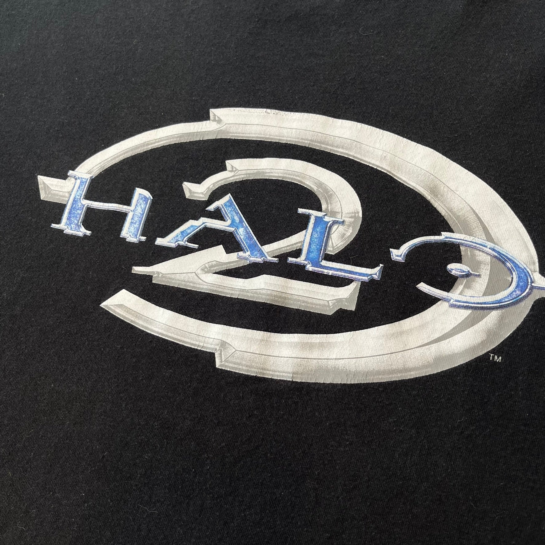 Halo 2 Xbox 2004 Black Tee is a black promotional T-shirt featuring a large white and blue Halo 2 graphic on the front. The upper back has a green original Xbox logo positioned below the collar.