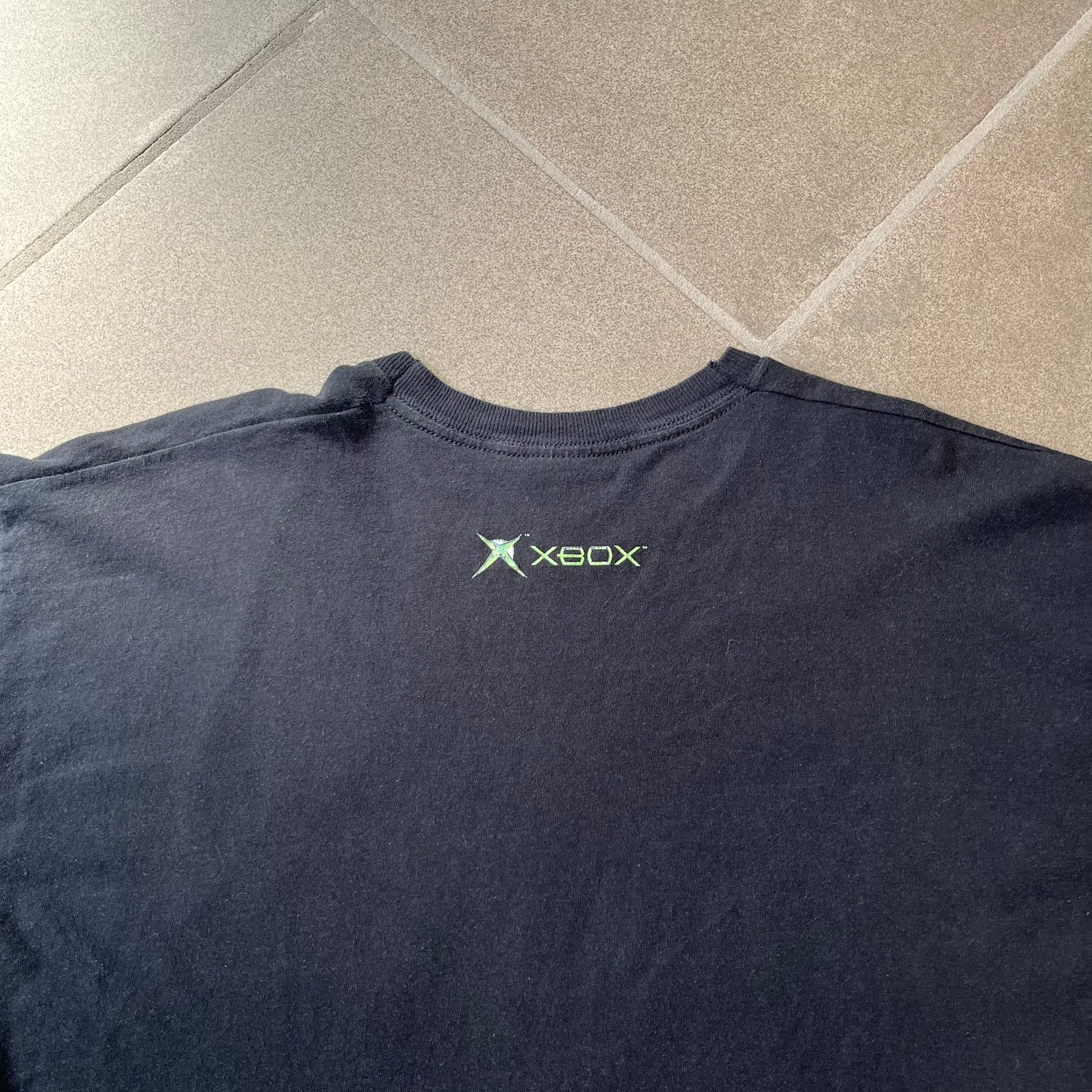 Halo 2 Xbox 2004 Black Tee is a black promotional T-shirt featuring a large white and blue Halo 2 graphic on the front. The upper back has a green original Xbox logo positioned below the collar.