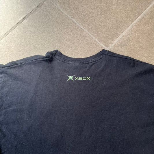 Halo 2 Xbox 2004 Black Tee is a black promotional T-shirt featuring a large white and blue Halo 2 graphic on the front. The upper back has a green original Xbox logo positioned below the collar.