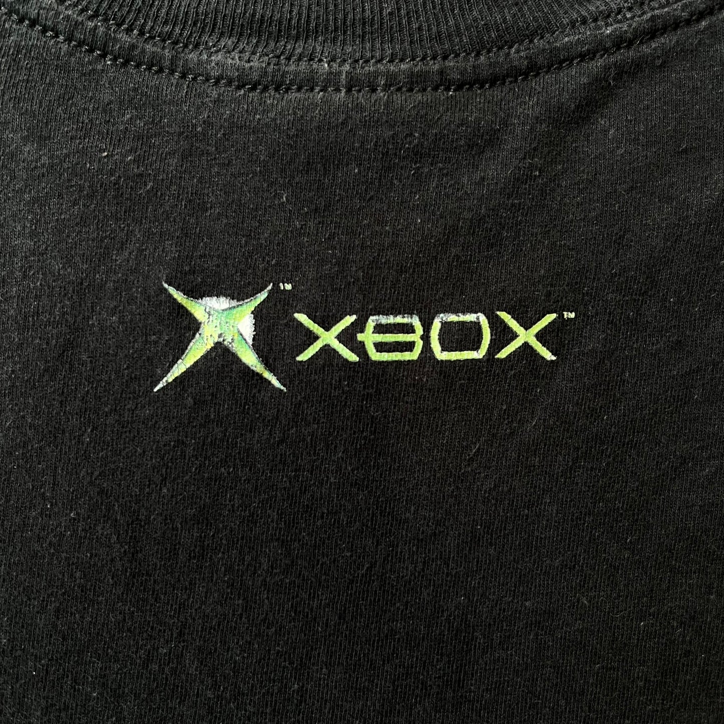 Halo 2 Xbox 2004 Black Tee is a black promotional T-shirt featuring a large white and blue Halo 2 graphic on the front. The upper back has a green original Xbox logo positioned below the collar.