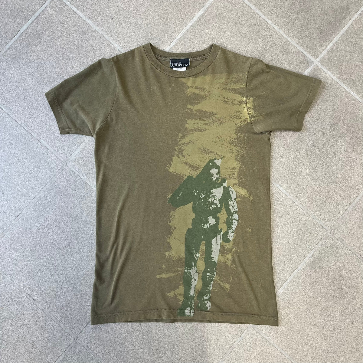 Halo 3 Finish the Fight 2006 Tee is a khaki, 100% cotton T-shirt with a green Master Chief graphic on the front against a lighter background. A Halo 3 logo is on one sleeve, and “Finish the Fight” text runs across the upper back. The neck tag is licensed for Xbox 360 and carries 2005 licensing on the other side.