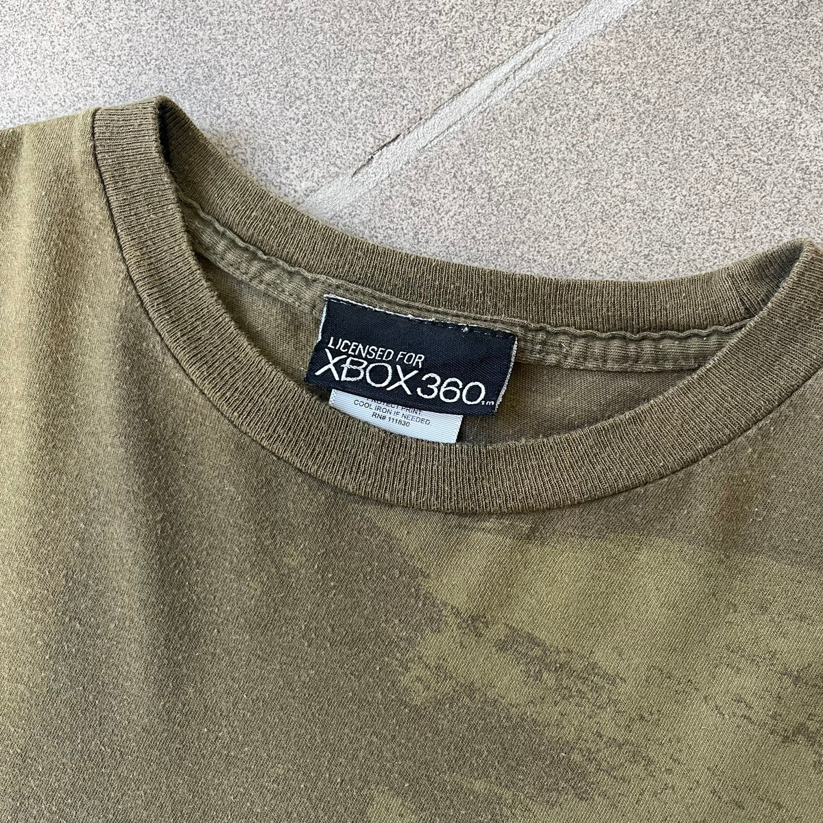 Halo 3 Finish the Fight 2006 Tee is a khaki, 100% cotton T-shirt with a green Master Chief graphic on the front against a lighter background. A Halo 3 logo is on one sleeve, and “Finish the Fight” text runs across the upper back. The neck tag is licensed for Xbox 360 and carries 2005 licensing on the other side.
