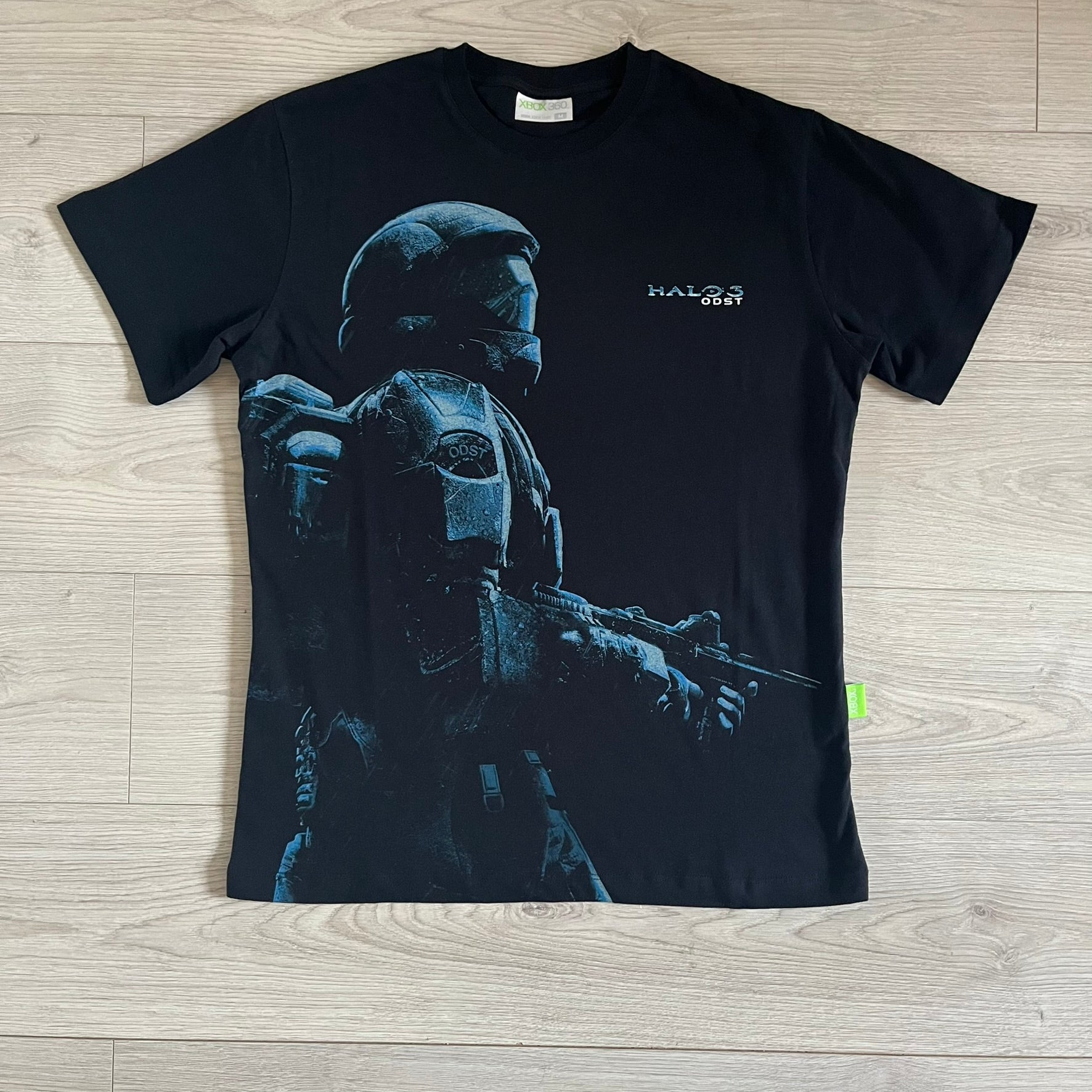 Black Halo 3: ODST Tee for Xbox 360 featuring a large blue Master Chief graphic on the front right and a small logo on the left chest. Made from high-quality cotton with Xbox 360 tags on the collar and a green one on the side. A super rare collectible for Halo fans.