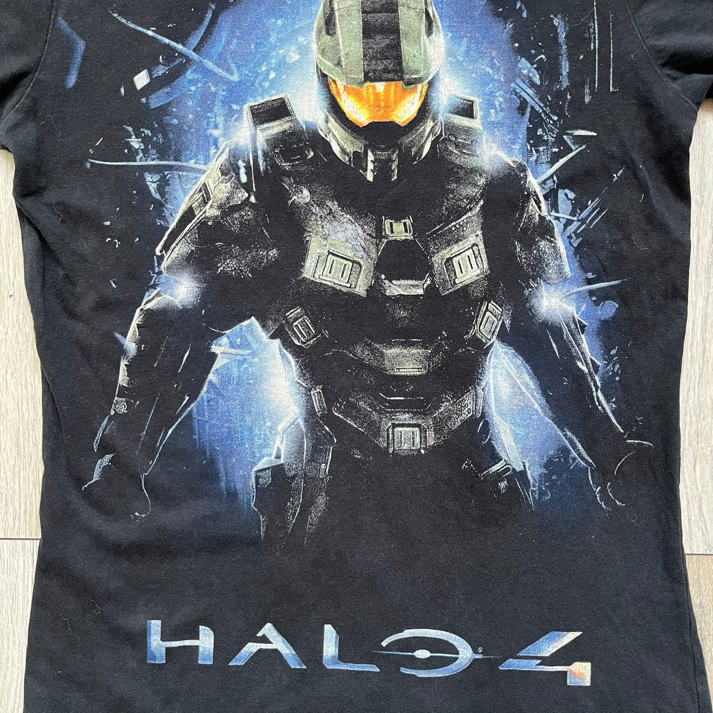 Halo 4 Master Chief 2012 Tee is a black graphic T-shirt featuring a large front print of Master Chief standing in front of a blue sci-fi style background. The design includes a detailed illustration of the character with a large Halo 4 logo printed below the graphic. The shirt has a 2012 Microsoft licensed neck tag.