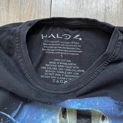 Halo 4 Master Chief 2012 Tee is a black graphic T-shirt featuring a large front print of Master Chief standing in front of a blue sci-fi style background. The design includes a detailed illustration of the character with a large Halo 4 logo printed below the graphic. The shirt has a 2012 Microsoft licensed neck tag.