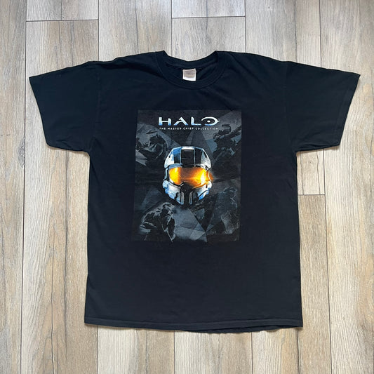 Halo The Master Chief Collection 2010s Tee is a black short-sleeve T-shirt featuring a front graphic of Master Chief’s helmet with golden-orange visor, surrounded by smaller Master Chief figures. The back displays white “@GAMEDIGITAL” text, referencing the gaming merchandise website. This T-shirt was produced as part of official Halo merchandise in the 2010s.