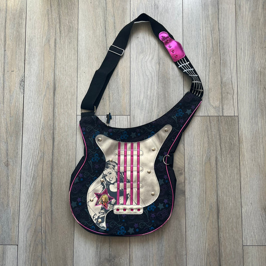 Hannah Montana 2000s Guitar-Shaped Shoulder Bag is a guitar-shaped bag with a black main body. The shoulder strap features pink glossy detailing and white string-like elements. The front panel has a multicolor pattern with stars, flowers, and butterflies. A large silver patch with pink string details and Hannah Montana graphics is placed on the front. The zipper pull is shaped with a Hannah Montana series logo design.
