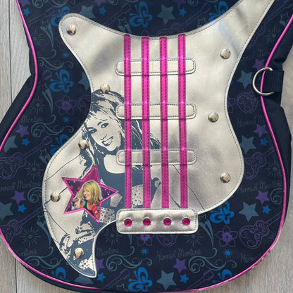 Hannah Montana 2000s Guitar-Shaped Shoulder Bag is a guitar-shaped bag with a black main body. The shoulder strap features pink glossy detailing and white string-like elements. The front panel has a multicolor pattern with stars, flowers, and butterflies. A large silver patch with pink string details and Hannah Montana graphics is placed on the front. The zipper pull is shaped with a Hannah Montana series logo design.
