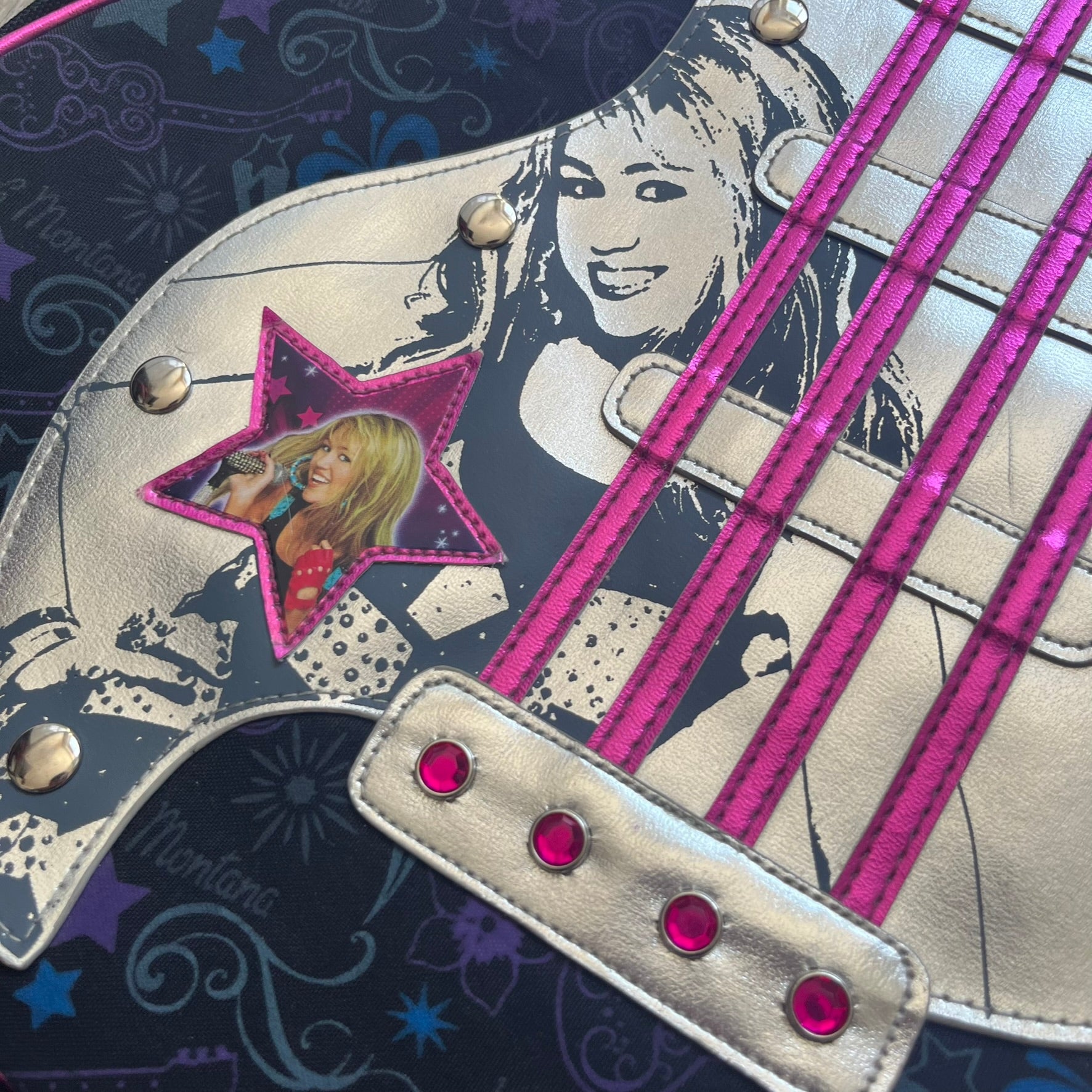Hannah Montana 2000s Guitar-Shaped Shoulder Bag is a guitar-shaped bag with a black main body. The shoulder strap features pink glossy detailing and white string-like elements. The front panel has a multicolor pattern with stars, flowers, and butterflies. A large silver patch with pink string details and Hannah Montana graphics is placed on the front. The zipper pull is shaped with a Hannah Montana series logo design.
