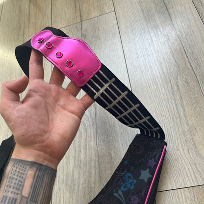 Hannah Montana 2000s Guitar-Shaped Shoulder Bag is a guitar-shaped bag with a black main body. The shoulder strap features pink glossy detailing and white string-like elements. The front panel has a multicolor pattern with stars, flowers, and butterflies. A large silver patch with pink string details and Hannah Montana graphics is placed on the front. The zipper pull is shaped with a Hannah Montana series logo design.
