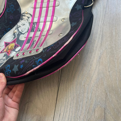Hannah Montana 2000s Guitar-Shaped Shoulder Bag is a guitar-shaped bag with a black main body. The shoulder strap features pink glossy detailing and white string-like elements. The front panel has a multicolor pattern with stars, flowers, and butterflies. A large silver patch with pink string details and Hannah Montana graphics is placed on the front. The zipper pull is shaped with a Hannah Montana series logo design.
