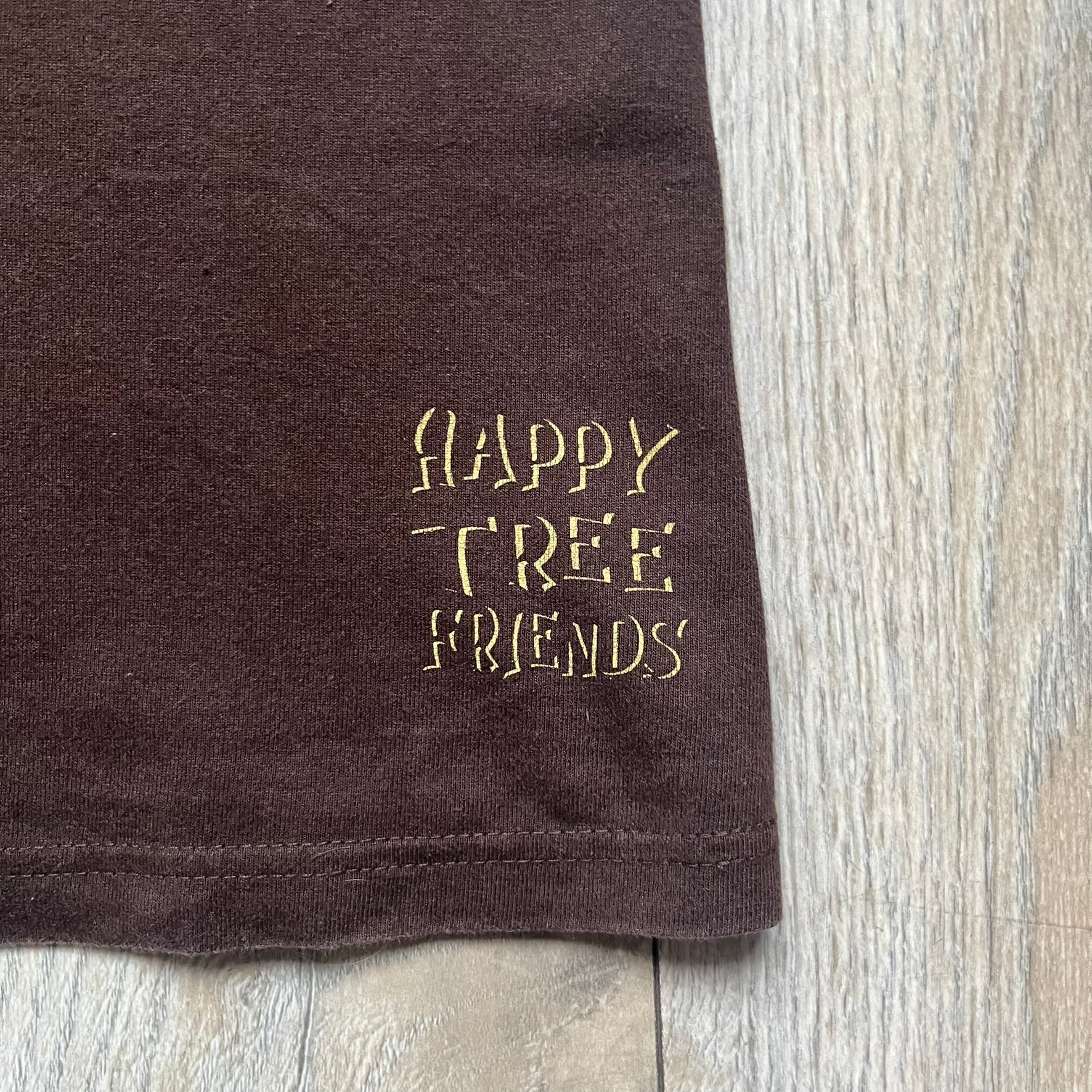 Happy Tree Friends Lumpy Fan Brown Tee from 2005 is a brown T-shirt featuring Lumpy on the front holding and licking a fan. On the back, the same Lumpy graphic appears without the tongue, with visible blood on the fan and face. A small Happy Tree Friends logo is printed in the lower front corner, and the neck tag features a red heart detail.