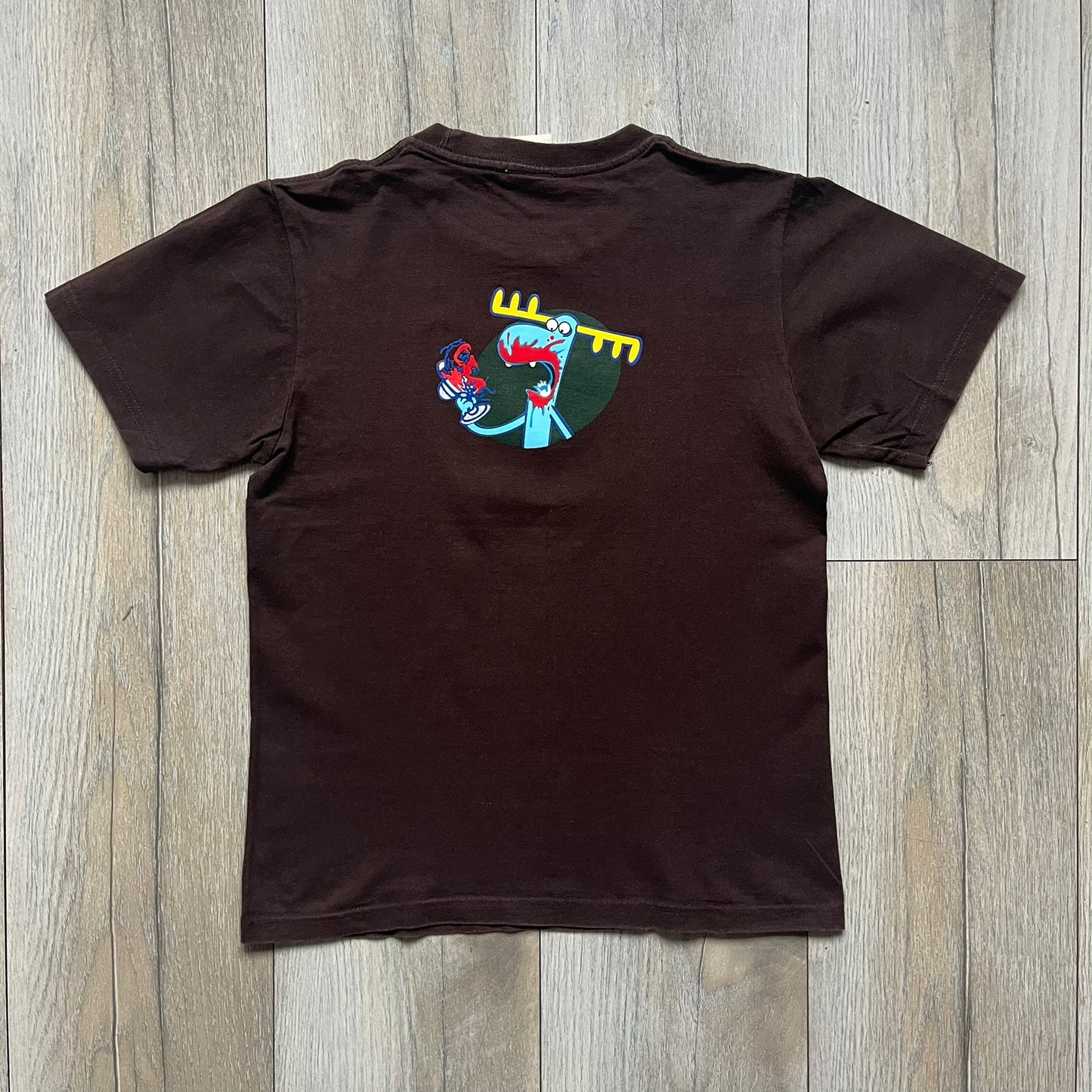 Happy Tree Friends Lumpy Fan Brown Tee from 2005 is a brown T-shirt featuring Lumpy on the front holding and licking a fan. On the back, the same Lumpy graphic appears without the tongue, with visible blood on the fan and face. A small Happy Tree Friends logo is printed in the lower front corner, and the neck tag features a red heart detail.