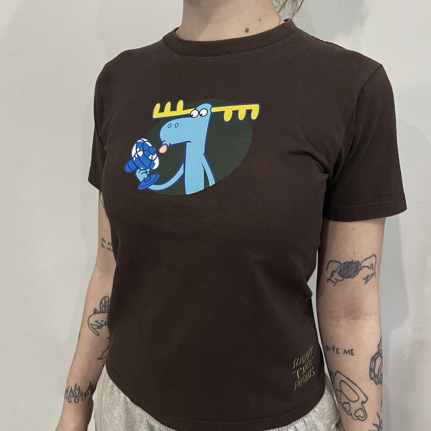 Happy Tree Friends Lumpy Fan Brown Tee from 2005 is a brown T-shirt featuring Lumpy on the front holding and licking a fan. On the back, the same Lumpy graphic appears without the tongue, with visible blood on the fan and face. A small Happy Tree Friends logo is printed in the lower front corner, and the neck tag features a red heart detail.