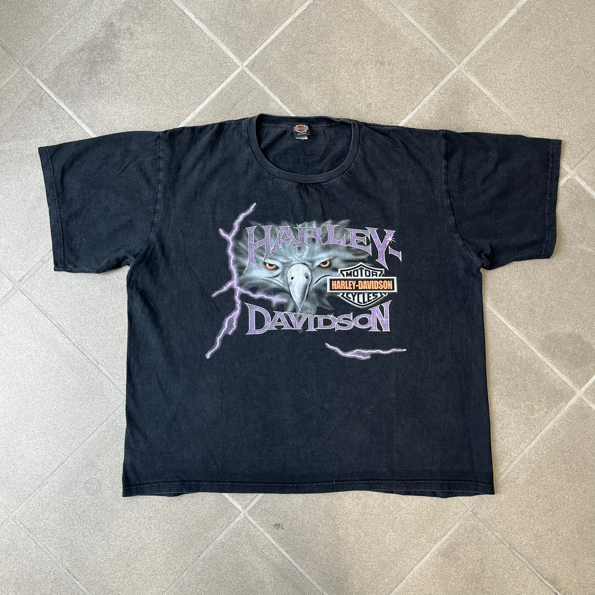 Harley-Davidson 2000's Eagle Black Tee features a boxy black T-shirt with a front graphic of an eagle's head alongside a Harley-Davidson logo. Purple and white “Harley Davidson” text and “Thunderstrikes” graphics surround the design. The back has a large white and grey graphic of a flying eagle with Harley-Davidson text underneath.
