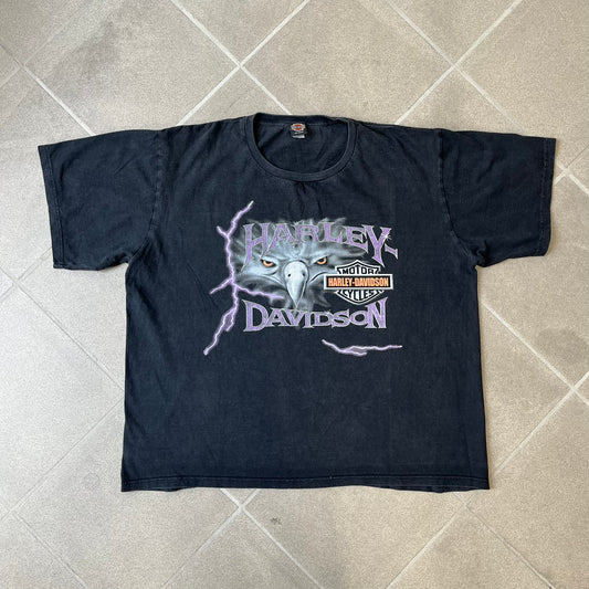 Harley-Davidson 2000's Eagle Black Tee features a boxy black T-shirt with a front graphic of an eagle's head alongside a Harley-Davidson logo. Purple and white “Harley Davidson” text and “Thunderstrikes” graphics surround the design. The back has a large white and grey graphic of a flying eagle with Harley-Davidson text underneath.
