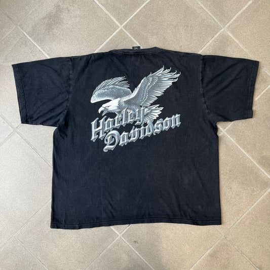 Harley-Davidson 2000's Eagle Black Tee features a boxy black T-shirt with a front graphic of an eagle's head alongside a Harley-Davidson logo. Purple and white “Harley Davidson” text and “Thunderstrikes” graphics surround the design. The back has a large white and grey graphic of a flying eagle with Harley-Davidson text underneath.
