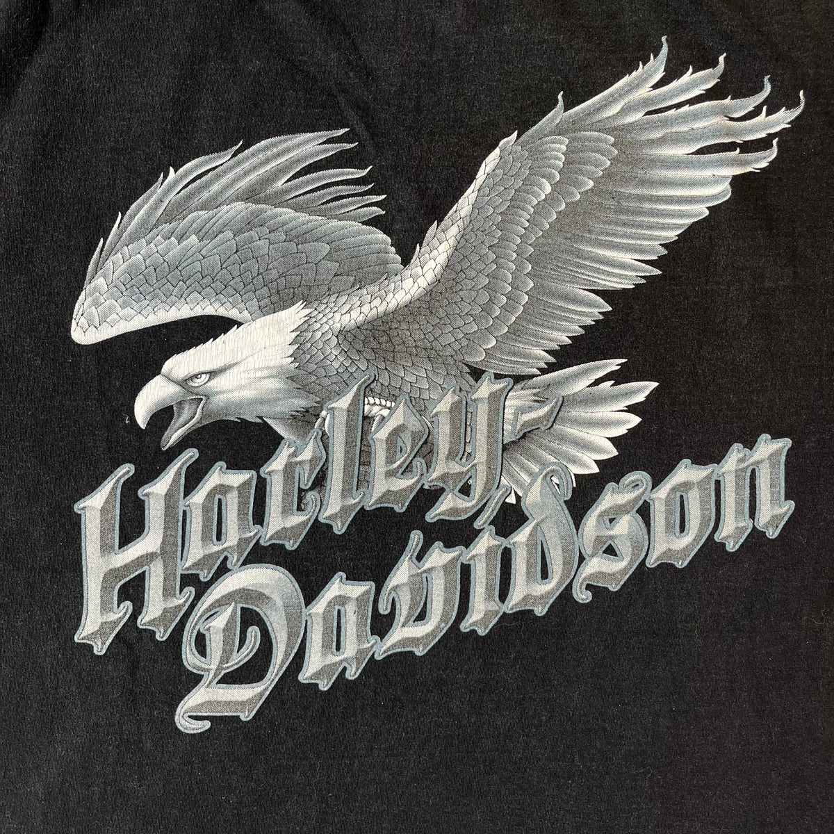 Harley-Davidson 2000's Eagle Black Tee features a boxy black T-shirt with a front graphic of an eagle's head alongside a Harley-Davidson logo. Purple and white “Harley Davidson” text and “Thunderstrikes” graphics surround the design. The back has a large white and grey graphic of a flying eagle with Harley-Davidson text underneath.
