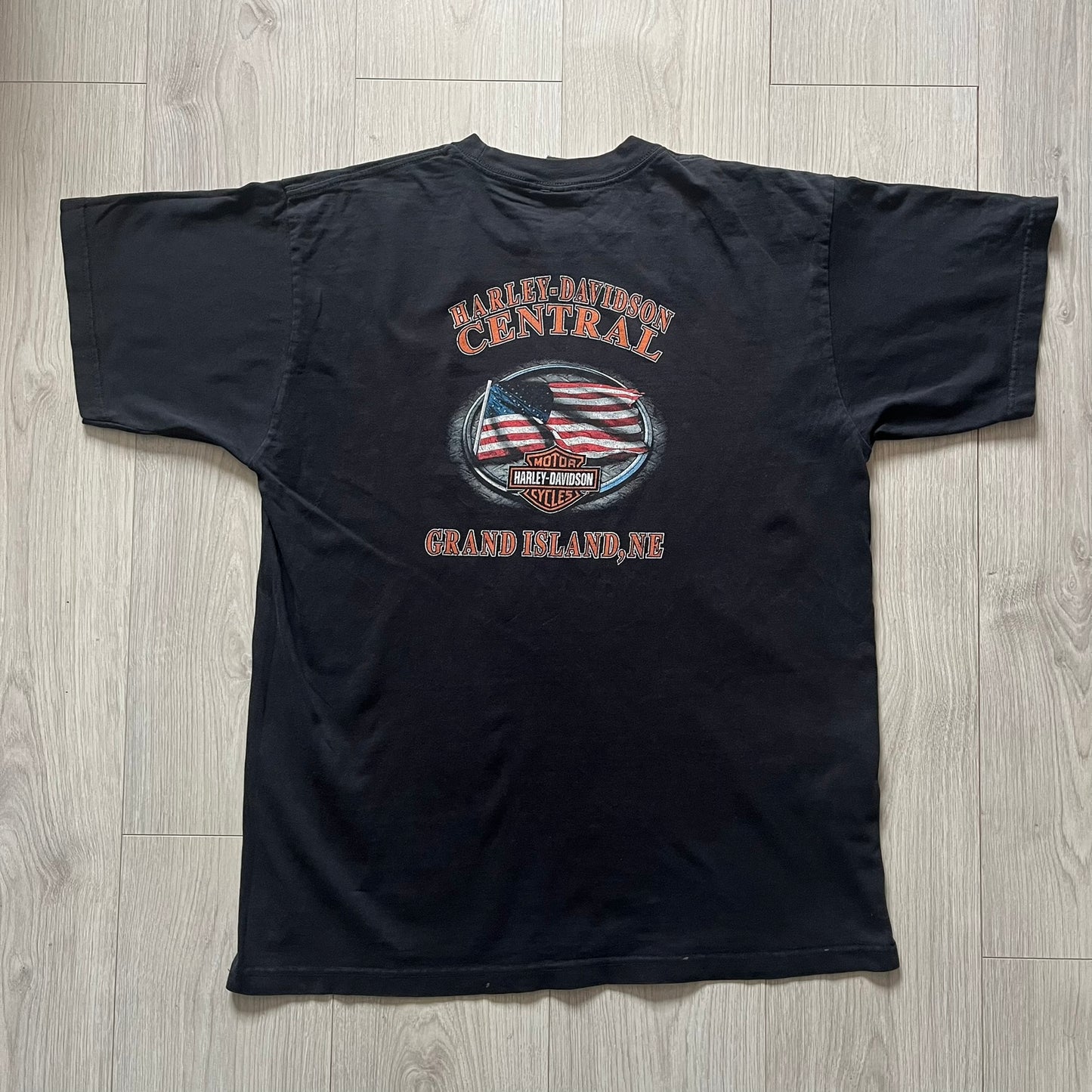  Harley Davidson Grand Island 2000’s Tee featuring a fiery Harley Davidson Motorcycles logo on the front and a USA flag graphic with Harley branding and “Grand Island, NE” text on the back. Classic black vintage tee with a timeless biker aesthetic.