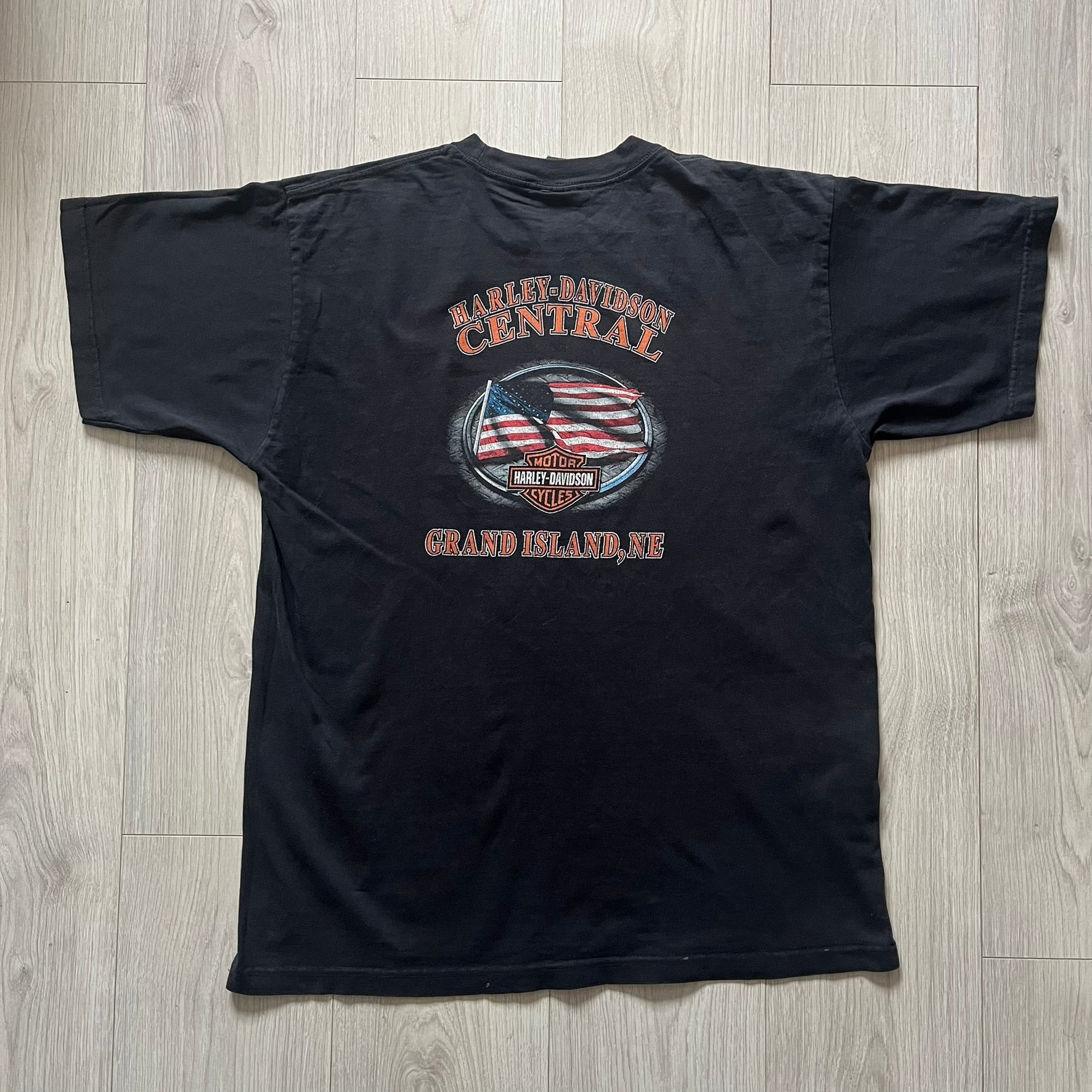 Harley Davidson Grand Island 2000’s Tee featuring a fiery Harley Davidson Motorcycles logo on the front and a USA flag graphic with Harley branding and “Grand Island, NE” text on the back. Classic black vintage tee with a timeless biker aesthetic.
