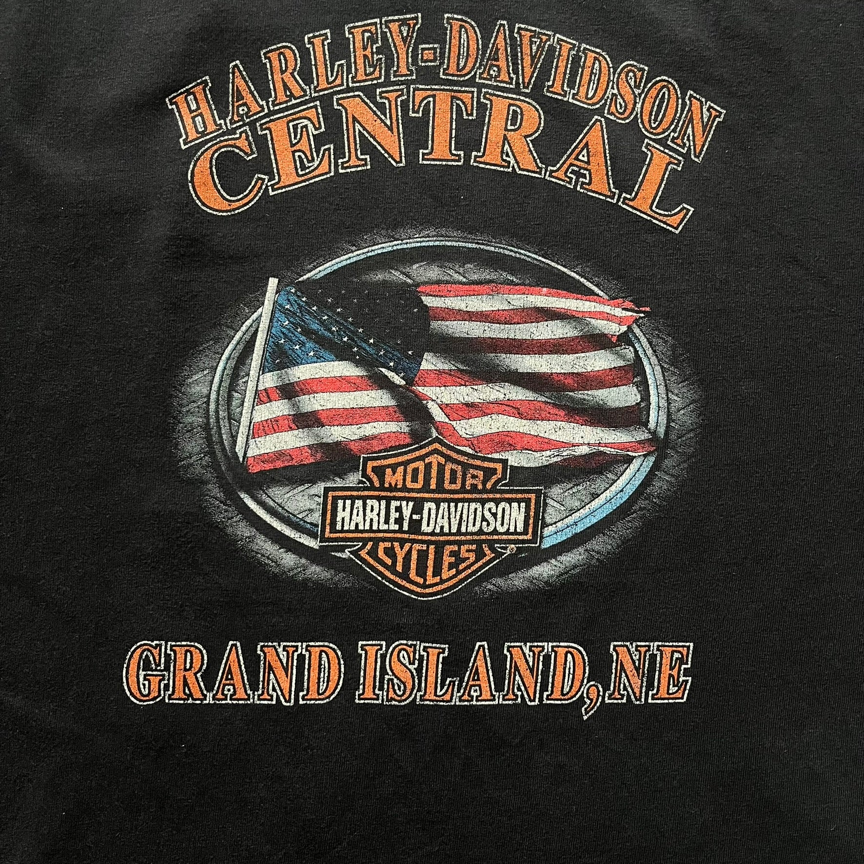 Harley Davidson Grand Island 2000’s Tee featuring a fiery Harley Davidson Motorcycles logo on the front and a USA flag graphic with Harley branding and “Grand Island, NE” text on the back. Classic black vintage tee with a timeless biker aesthetic.