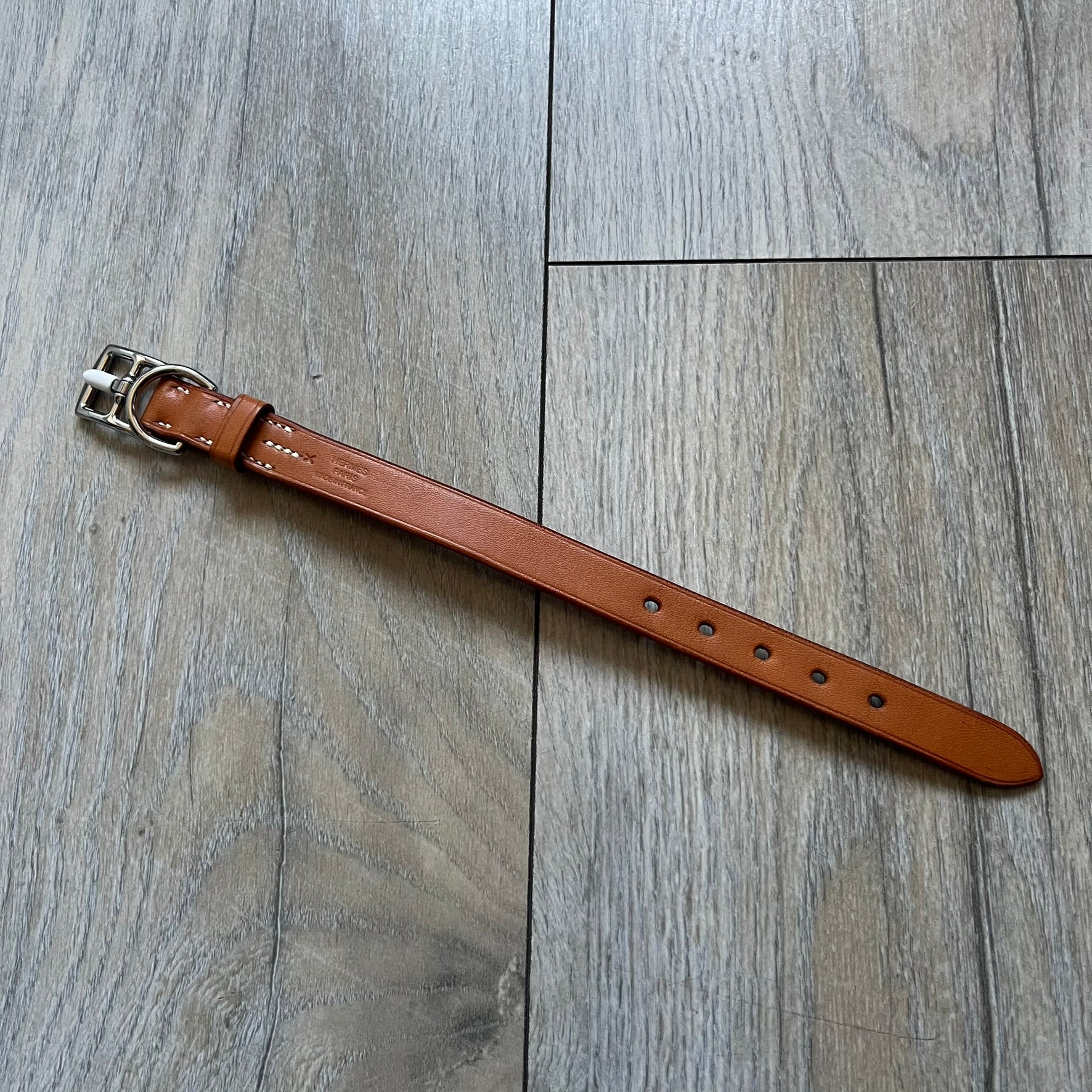 Small model Hermès Étrivière dog collar crafted from Natural Sable bridle leather. The collar features a stirrup-shaped buckle stamped HERMÈS, a metal D-ring for leash attachment, and leather stamped “HERMÈS PARIS MADE IN FRANCE.” It includes a 2 cm silver-plated stainless steel medal engraved “Hermès Sellier Paris,” with the reverse side suitable for engraving