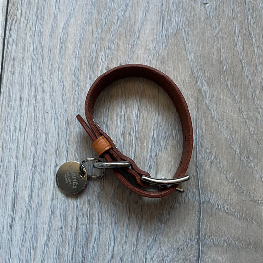 Small model Hermès Étrivière dog collar crafted from Natural Sable bridle leather. The collar features a stirrup-shaped buckle stamped HERMÈS, a metal D-ring for leash attachment, and leather stamped “HERMÈS PARIS MADE IN FRANCE.” It includes a 2 cm silver-plated stainless steel medal engraved “Hermès Sellier Paris,” with the reverse side suitable for engraving