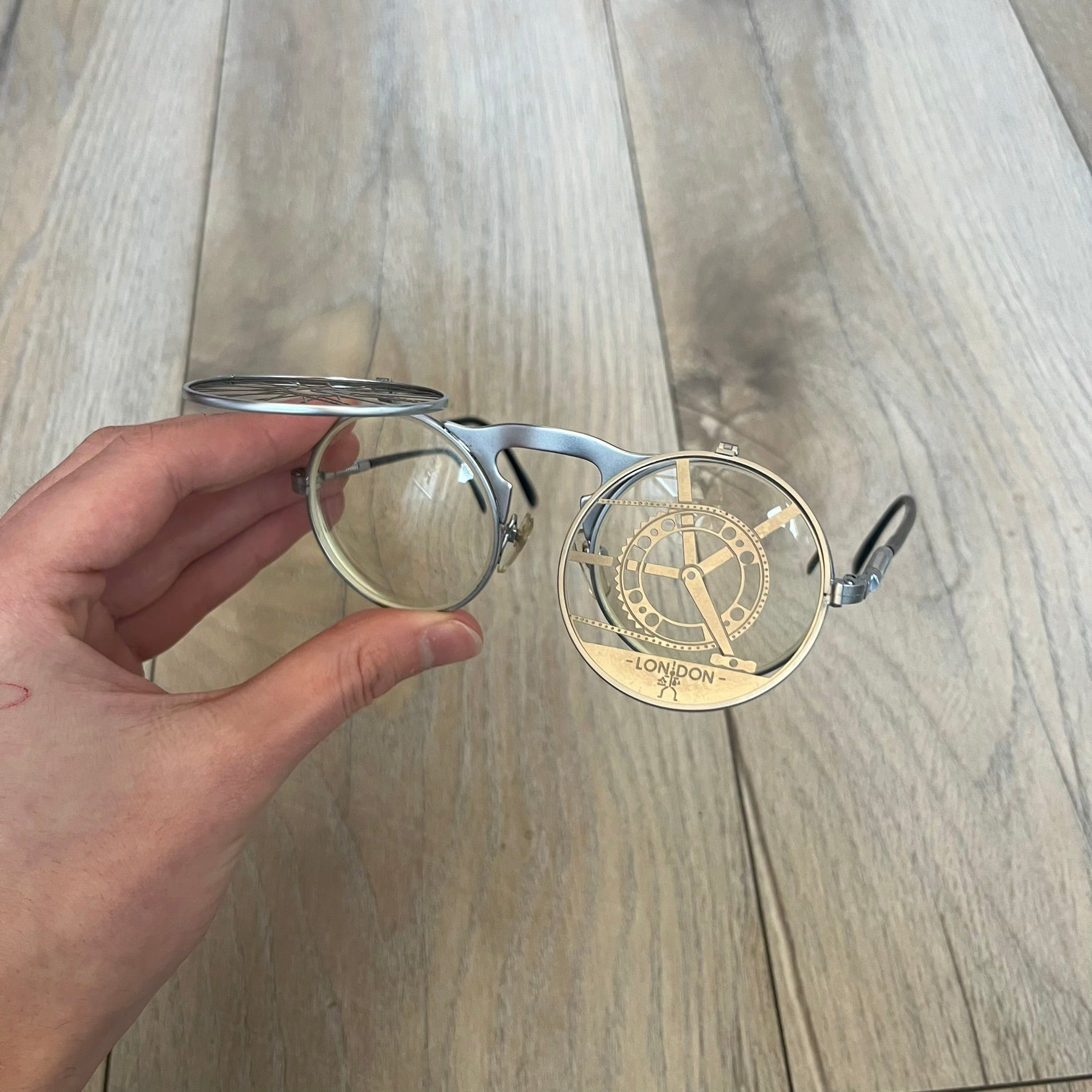 Hi-Tek Alexander London HT-002 2000s Flip-Up Metal Glasses are prescription eyeglasses featuring metal flip-up covers designed as bicycle wheels with visible spokes and pedals, forming a bicycle shape across the frame when closed. One flap is detailed with a Hi-Tek Alexander London logo, while the frame includes black temple arms. The model comes with a black leather and metal case, and the lenses are prescription, requiring replacement for use.