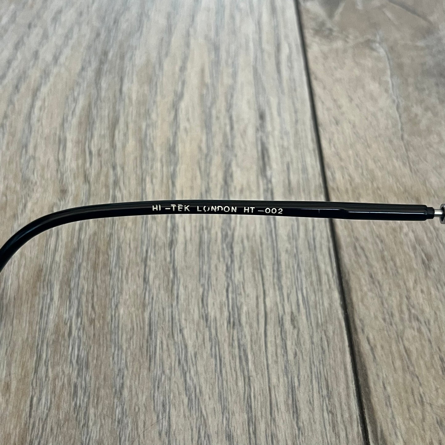 Hi-Tek Alexander London HT-002 2000s Flip-Up Metal Glasses are prescription eyeglasses featuring metal flip-up covers designed as bicycle wheels with visible spokes and pedals, forming a bicycle shape across the frame when closed. One flap is detailed with a Hi-Tek Alexander London logo, while the frame includes black temple arms. The model comes with a black leather and metal case, and the lenses are prescription, requiring replacement for use.