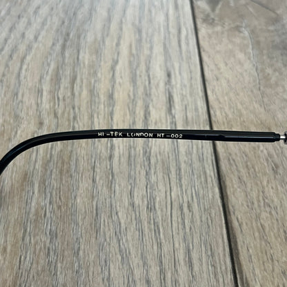 Hi-Tek Alexander London HT-002 2000s Flip-Up Metal Glasses are prescription eyeglasses featuring metal flip-up covers designed as bicycle wheels with visible spokes and pedals, forming a bicycle shape across the frame when closed. One flap is detailed with a Hi-Tek Alexander London logo, while the frame includes black temple arms. The model comes with a black leather and metal case, and the lenses are prescription, requiring replacement for use.