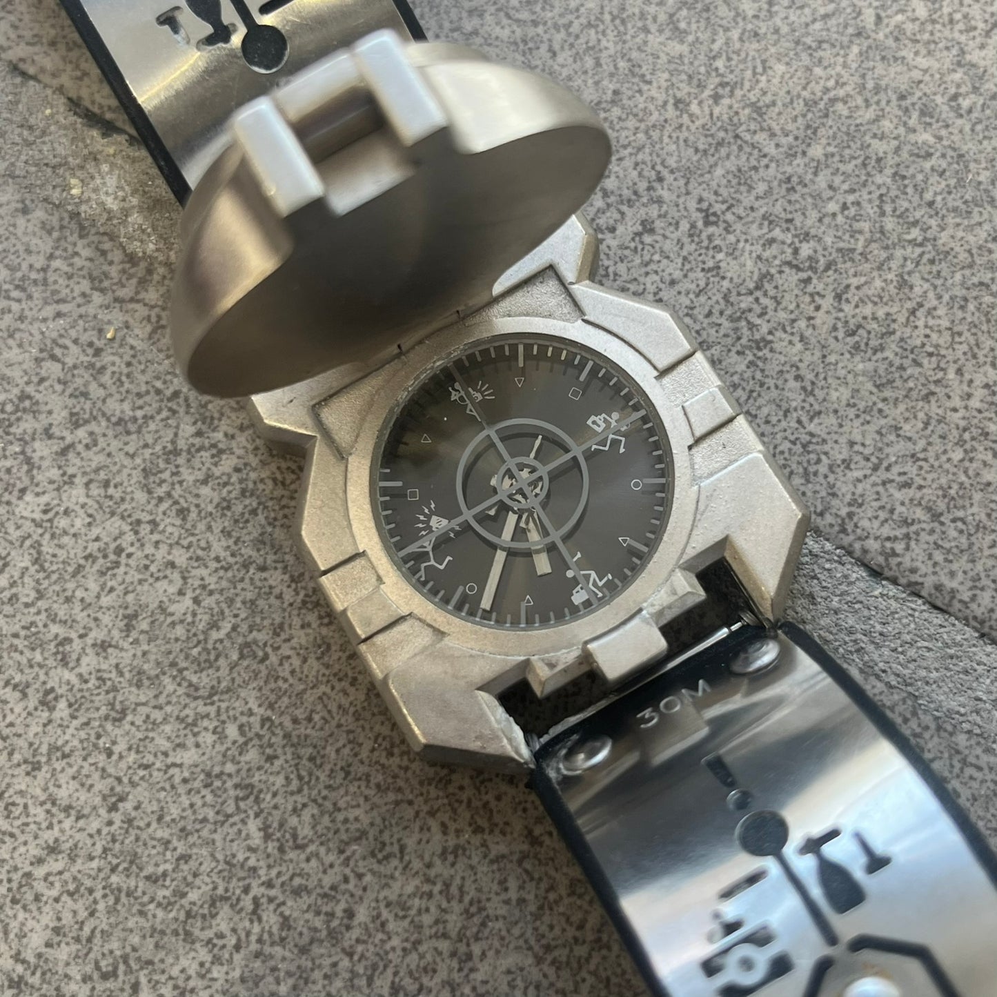 Hi-Tek Alexander 90s Crosshair Double Latch Analog Watch features a unique dual-latch case design. The first metal latch fully covers the dial, while the second thicker latch includes a hidden cavity-like space. The black dial features Hi-Tek figure markers instead of traditional hours, with a grey crosshair detail across the face. The watch has rigid metal straps with Hi-Tek Designs branding and “Made in England” markings, while the inner side of the bracelet has a black finish. The bracelet uses an unusua