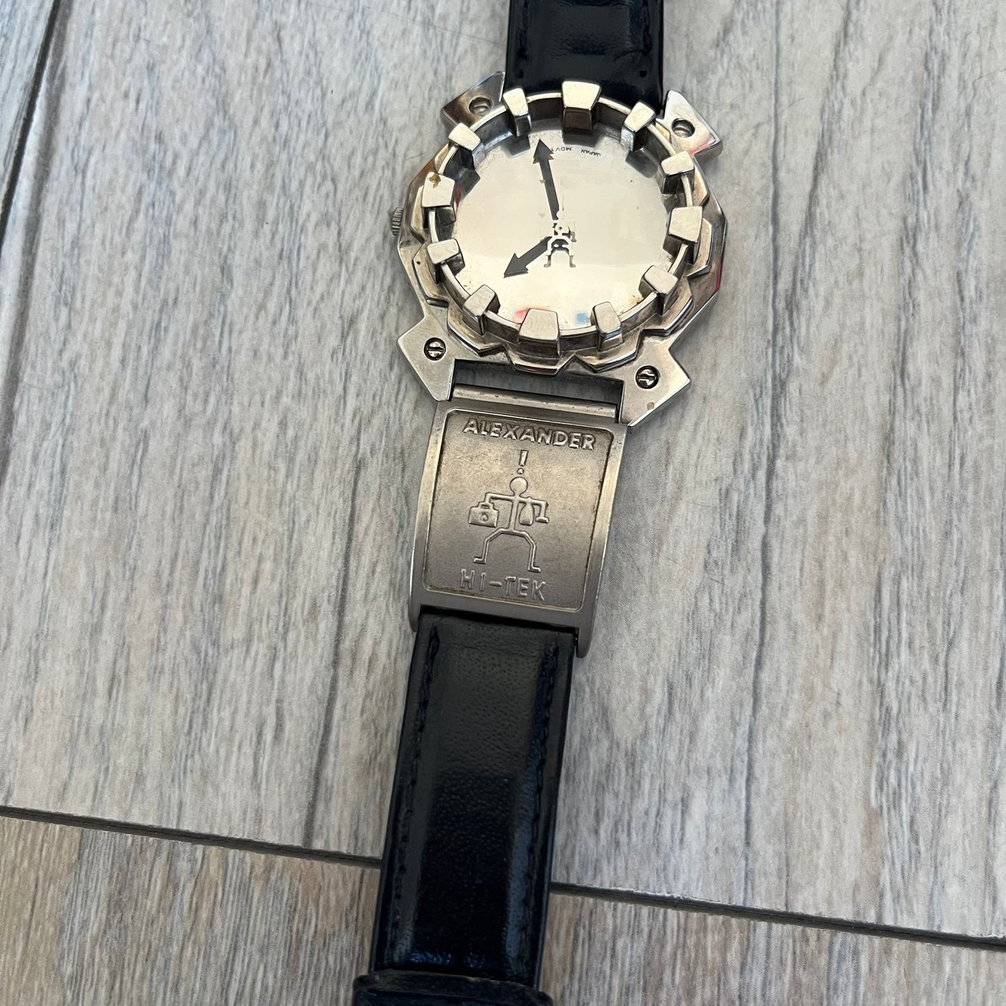 Hi-Tek Alexander 90s Mirror Cavity Analog Watch is a Hi-Tek Alexander watch with black leather straps. One strap features a metal Hi-Tek Alexander plaque with the brand figure. The dial has a mirrored metallic finish with raised square markers instead of traditional hour indices. The hands are black, and the seconds hand is designed as a Hi-Tek figure.
