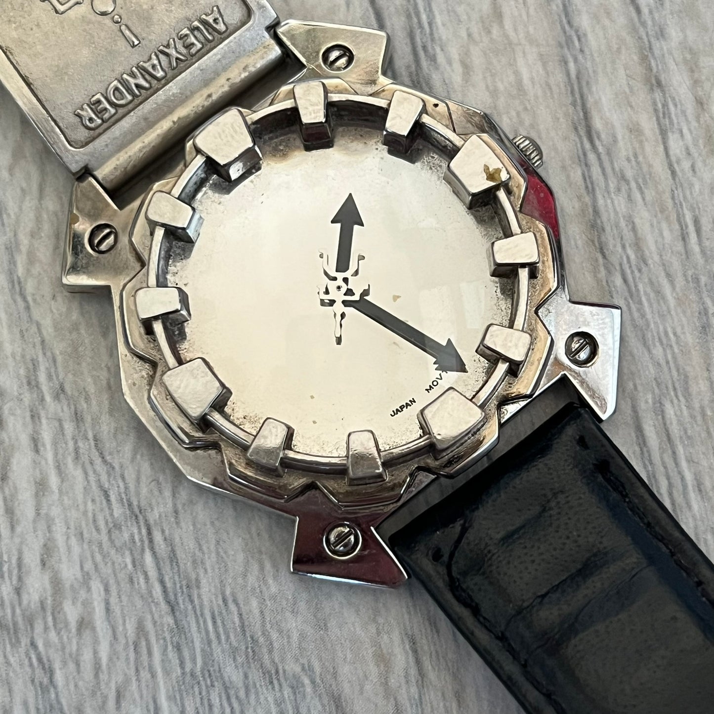 Hi-Tek Alexander 90s Mirror Cavity Analog Watch is a Hi-Tek Alexander watch with black leather straps. One strap features a metal Hi-Tek Alexander plaque with the brand figure. The dial has a mirrored metallic finish with raised square markers instead of traditional hour indices. The hands are black, and the seconds hand is designed as a Hi-Tek figure.
