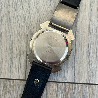 Hi-Tek Alexander 90s Mirror Cavity Analog Watch is a Hi-Tek Alexander watch with black leather straps. One strap features a metal Hi-Tek Alexander plaque with the brand figure. The dial has a mirrored metallic finish with raised square markers instead of traditional hour indices. The hands are black, and the seconds hand is designed as a Hi-Tek figure.
