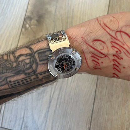 Hi-Tek Alexander 90s Perforated Analog Watch is a Hi-Tek London watch featuring a black dial with orange hands. The hour markers are represented by Hi-Tek figure graphics instead of traditional numerals. The dial is covered by a metal protective plate with a perforated hole pattern. Around the case is a metal ring with Hi-Tek London branding and a figure detail. The bracelet consists of two rigid metal sections with alternating small and large perforations.