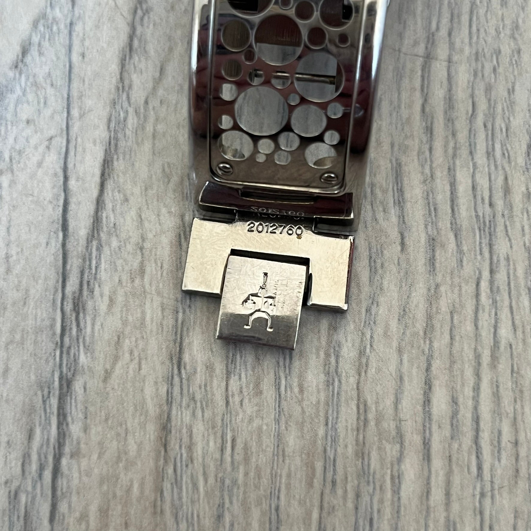 Hi-Tek Alexander 90s Perforated Analog Watch is a Hi-Tek London watch featuring a black dial with orange hands. The hour markers are represented by Hi-Tek figure graphics instead of traditional numerals. The dial is covered by a metal protective plate with a perforated hole pattern. Around the case is a metal ring with Hi-Tek London branding and a figure detail. The bracelet consists of two rigid metal sections with alternating small and large perforations.
