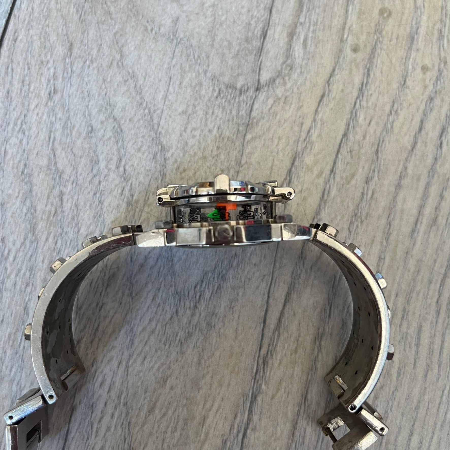 Hi-Tek Alexander 90's Side View Latch Analog Watch is a Hi-Tek Alexander watch with a rigid metal bracelet made of two sections featuring a raised circular pattern. The watch case has a latch-style cover that can be closed over the dial and is fully metallic. The dial is black and replaces traditional numbers with Hi-Tek figures - seconds hand is also a figure, while the hour and minute indicators are colored green and orange. The sides of the case are made of glass, allowing the time to be viewed from mult