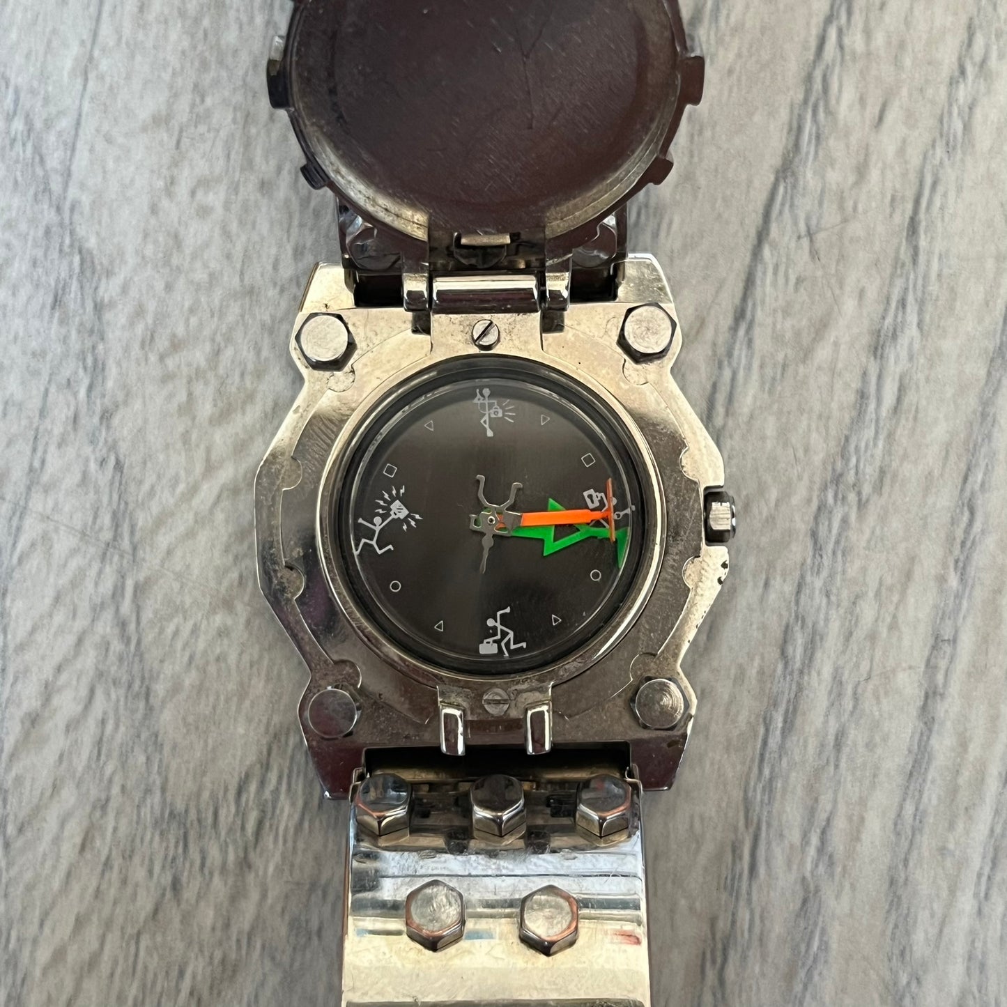 Hi-Tek Alexander 90's Side View Latch Analog Watch is a Hi-Tek Alexander watch with a rigid metal bracelet made of two sections featuring a raised circular pattern. The watch case has a latch-style cover that can be closed over the dial and is fully metallic. The dial is black and replaces traditional numbers with Hi-Tek figures - seconds hand is also a figure, while the hour and minute indicators are colored green and orange. The sides of the case are made of glass, allowing the time to be viewed from mult