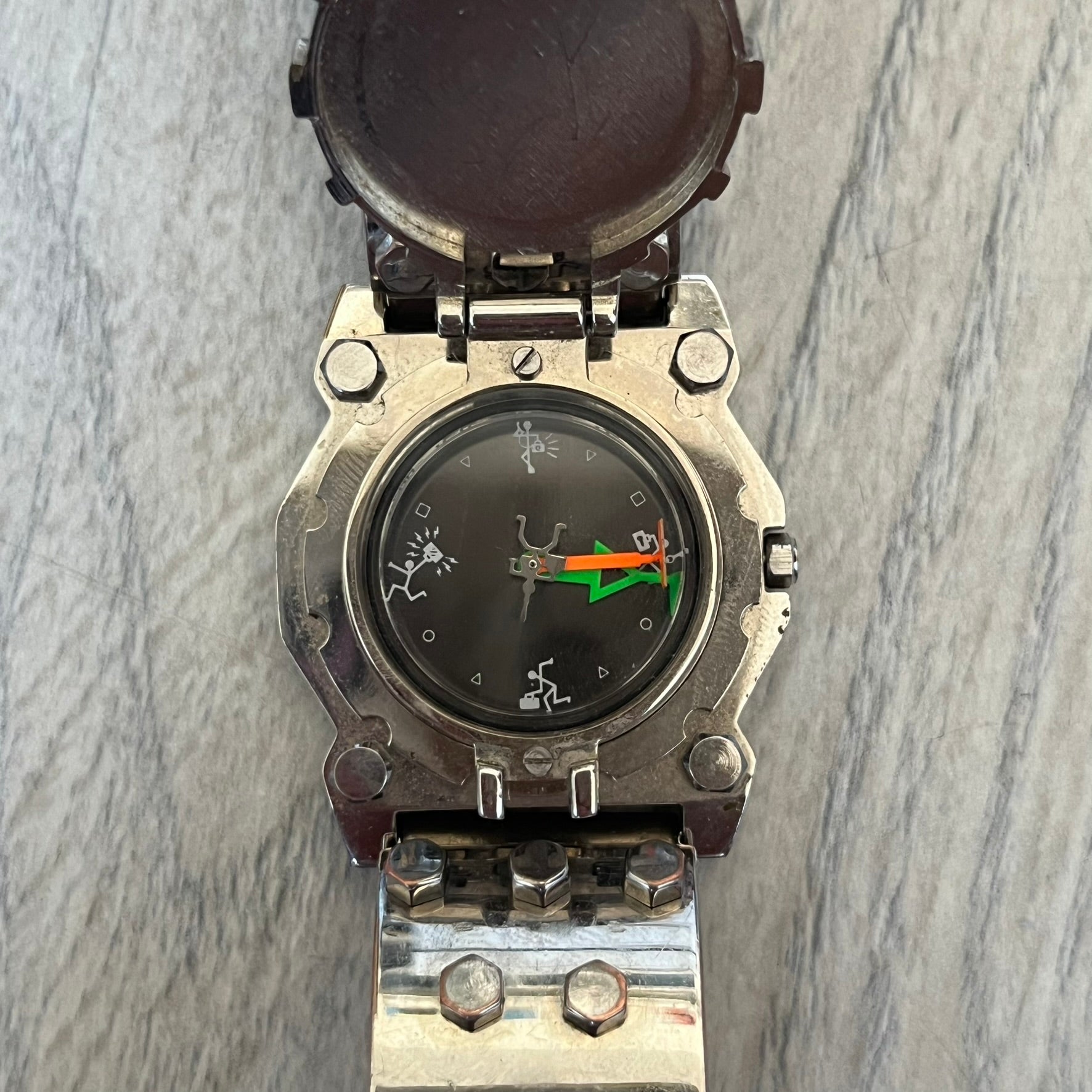 Hi-Tek Alexander 90's Side View Latch Analog Watch is a Hi-Tek Alexander watch with a rigid metal bracelet made of two sections featuring a raised circular pattern. The watch case has a latch-style cover that can be closed over the dial and is fully metallic. The dial is black and replaces traditional numbers with Hi-Tek figures - seconds hand is also a figure, while the hour and minute indicators are colored green and orange. The sides of the case are made of glass, allowing the time to be viewed from mult
