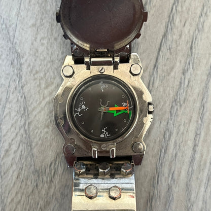 Hi-Tek Alexander 90's Side View Latch Analog Watch is a Hi-Tek Alexander watch with a rigid metal bracelet made of two sections featuring a raised circular pattern. The watch case has a latch-style cover that can be closed over the dial and is fully metallic. The dial is black and replaces traditional numbers with Hi-Tek figures - seconds hand is also a figure, while the hour and minute indicators are colored green and orange. The sides of the case are made of glass, allowing the time to be viewed from mult