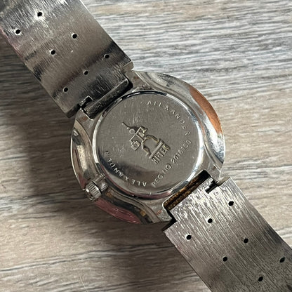 Hi-Tek Alexander “Cylinder” 90’s Analog Watch is a 1990s English-made timepiece featuring a black dial with a small Hi-Tek logo as the seconds hand. The cylindrical 3D case rises from the wrist, making the dial visible only from above, and the straps feature raised dot detailing. The stainless steel case back is branded with the Hi-Tek logo, designed specifically for smaller wrists.