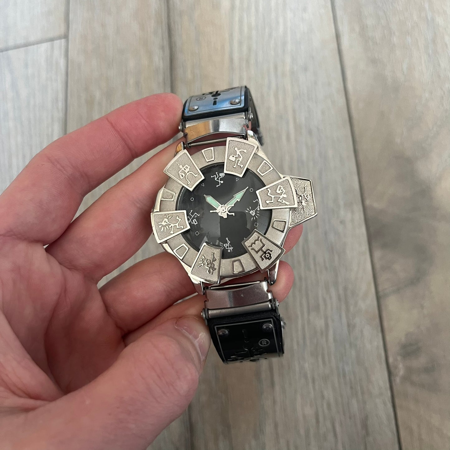 Hi-Tek Alexander “Running Man” 1990s Analog Watch is a stainless steel watch featuring a black dial and full steel case with integrated bracelet strap. The bezel includes a rotating top layer decorated with Hi-Tek character figures in various poses, while the dial replaces traditional numerals with similar character graphics. The seconds hand is designed as a character figure, and the hour and minute hands feature glow-in-the-dark functionality.