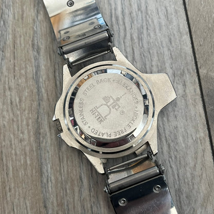 Hi-Tek Alexander “Running Man” 1990s Analog Watch is a stainless steel watch featuring a black dial and full steel case with integrated bracelet strap. The bezel includes a rotating top layer decorated with Hi-Tek character figures in various poses, while the dial replaces traditional numerals with similar character graphics. The seconds hand is designed as a character figure, and the hour and minute hands feature glow-in-the-dark functionality.