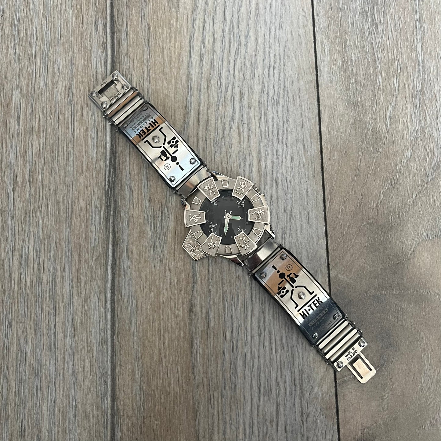 Hi-Tek Alexander “Running Man” 1990s Analog Watch is a stainless steel watch featuring a black dial and full steel case with integrated bracelet strap. The bezel includes a rotating top layer decorated with Hi-Tek character figures in various poses, while the dial replaces traditional numerals with similar character graphics. The seconds hand is designed as a character figure, and the hour and minute hands feature glow-in-the-dark functionality.