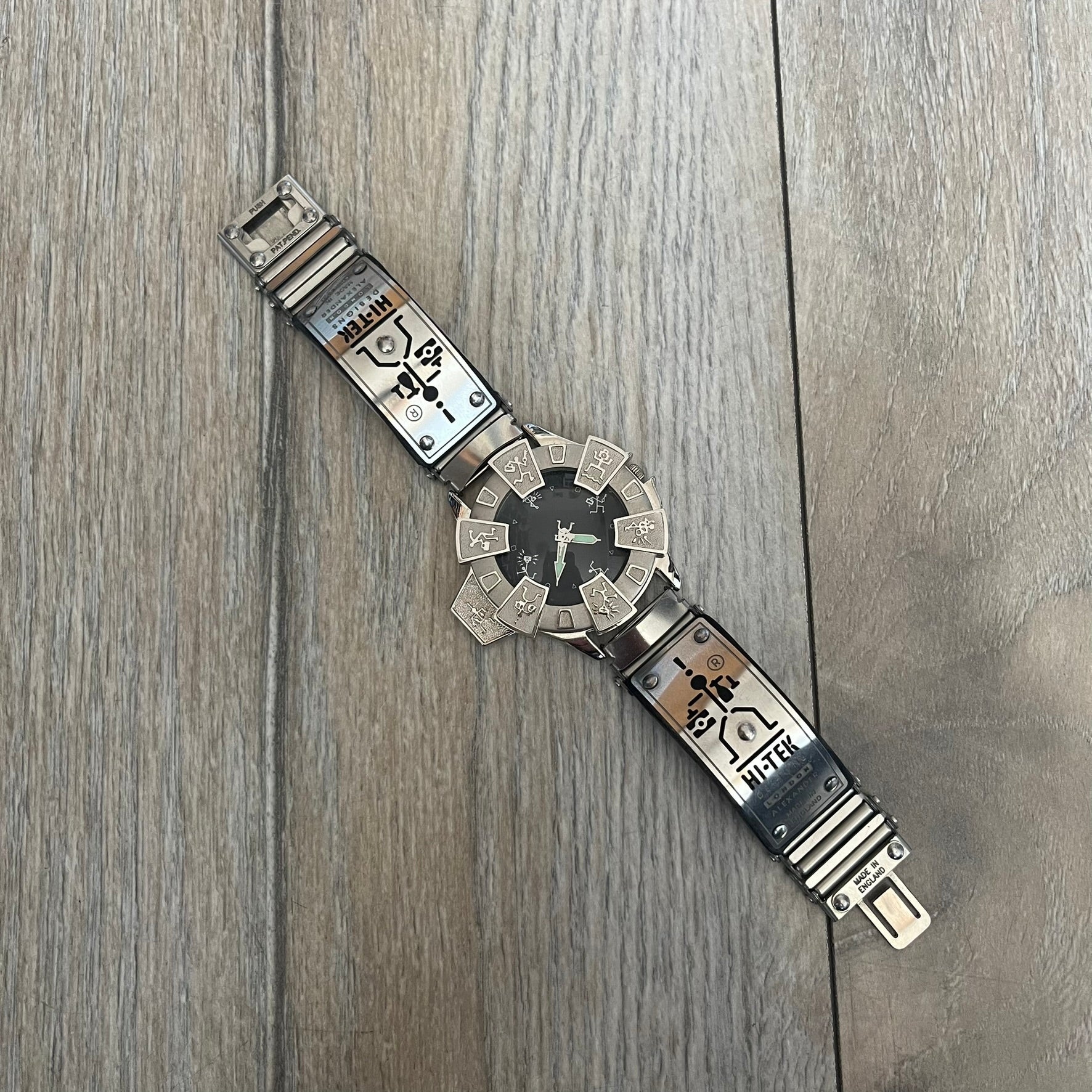 Hi-Tek Alexander “Running Man” 1990s Analog Watch is a stainless steel watch featuring a black dial and full steel case with integrated bracelet strap. The bezel includes a rotating top layer decorated with Hi-Tek character figures in various poses, while the dial replaces traditional numerals with similar character graphics. The seconds hand is designed as a character figure, and the hour and minute hands feature glow-in-the-dark functionality.
