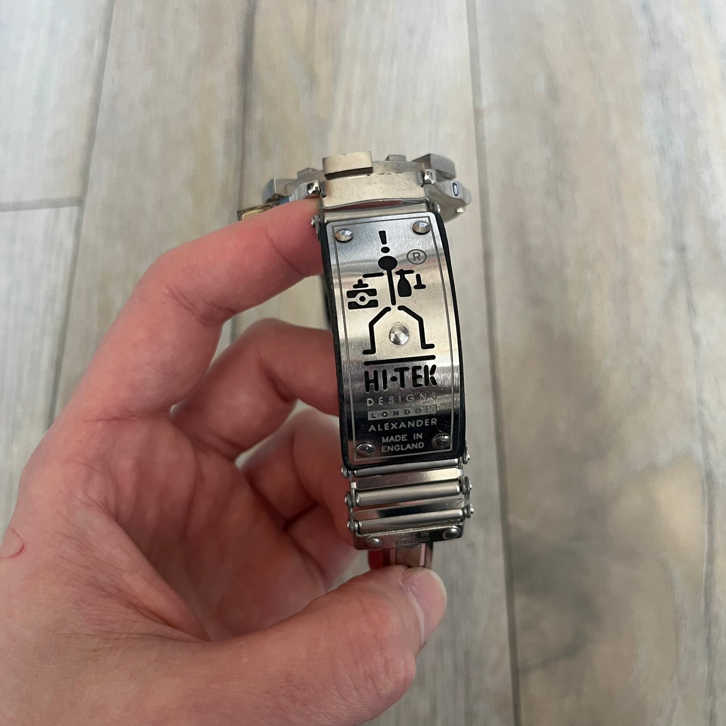 Hi-Tek Alexander “Running Man” 1990s Analog Watch is a stainless steel watch featuring a black dial and full steel case with integrated bracelet strap. The bezel includes a rotating top layer decorated with Hi-Tek character figures in various poses, while the dial replaces traditional numerals with similar character graphics. The seconds hand is designed as a character figure, and the hour and minute hands feature glow-in-the-dark functionality.