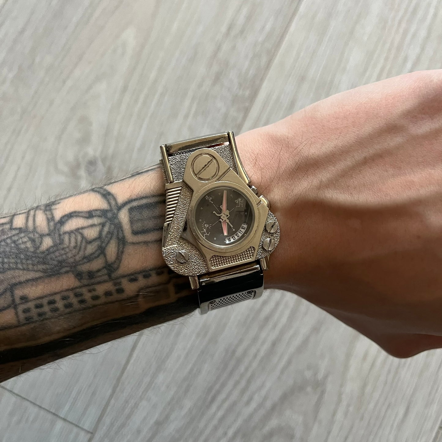 The Hi-Tek Alexander “The Engine” analog watch is a 1990s model made in England, designed with a sculptural case shaped like a motorcycle engine. It features a black dial with Alexander Hi-Tek figure logos in place of hour markers, pink hour and minute hands, and a Hi-Tek figure logo used as the seconds hand. A small sub-section at the bottom displays the day in numeric form, while visible screws and textured detailing are integrated into the engine-style face.&nbsp;The watch has an alloy case with a stainl