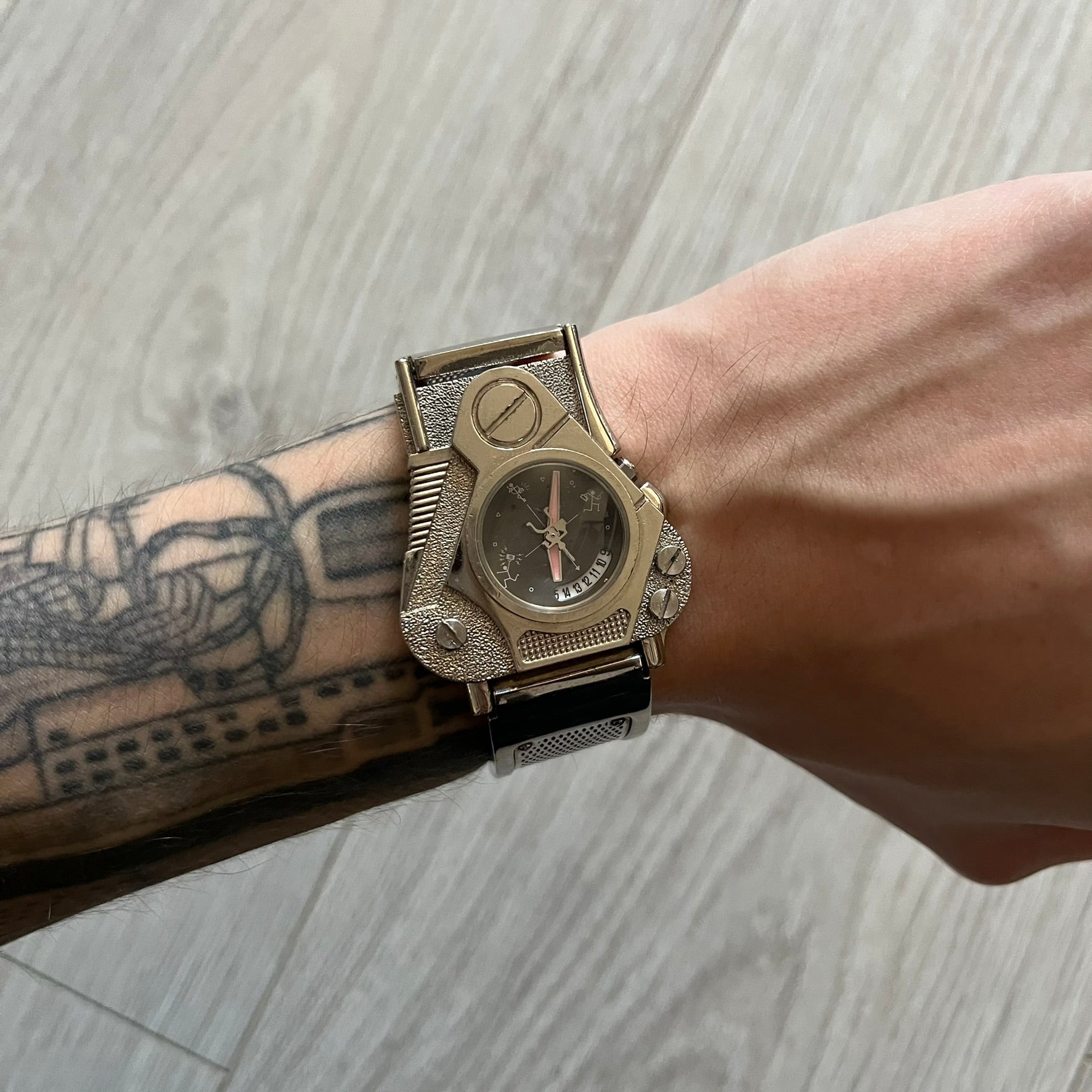 The Hi-Tek Alexander “The Engine” analog watch is a 1990s model made in England, designed with a sculptural case shaped like a motorcycle engine. It features a black dial with Alexander Hi-Tek figure logos in place of hour markers, pink hour and minute hands, and a Hi-Tek figure logo used as the seconds hand. A small sub-section at the bottom displays the day in numeric form, while visible screws and textured detailing are integrated into the engine-style face.&nbsp;The watch has an alloy case with a stainl