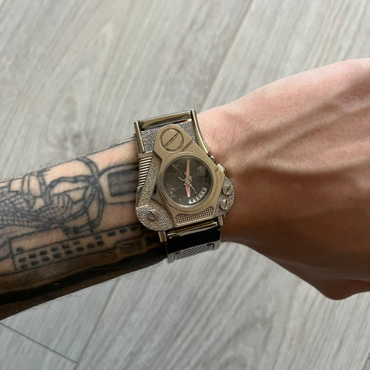 The Hi-Tek Alexander “The Engine” analog watch is a 1990s model made in England, designed with a sculptural case shaped like a motorcycle engine. It features a black dial with Alexander Hi-Tek figure logos in place of hour markers, pink hour and minute hands, and a Hi-Tek figure logo used as the seconds hand. A small sub-section at the bottom displays the day in numeric form, while visible screws and textured detailing are integrated into the engine-style face.&nbsp;The watch has an alloy case with a stainl